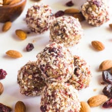 Several cherry vanilla energy bites are stacked. They are surrounded by raw almonds. There are raw almonds and dried cherries in a bowl behind it.