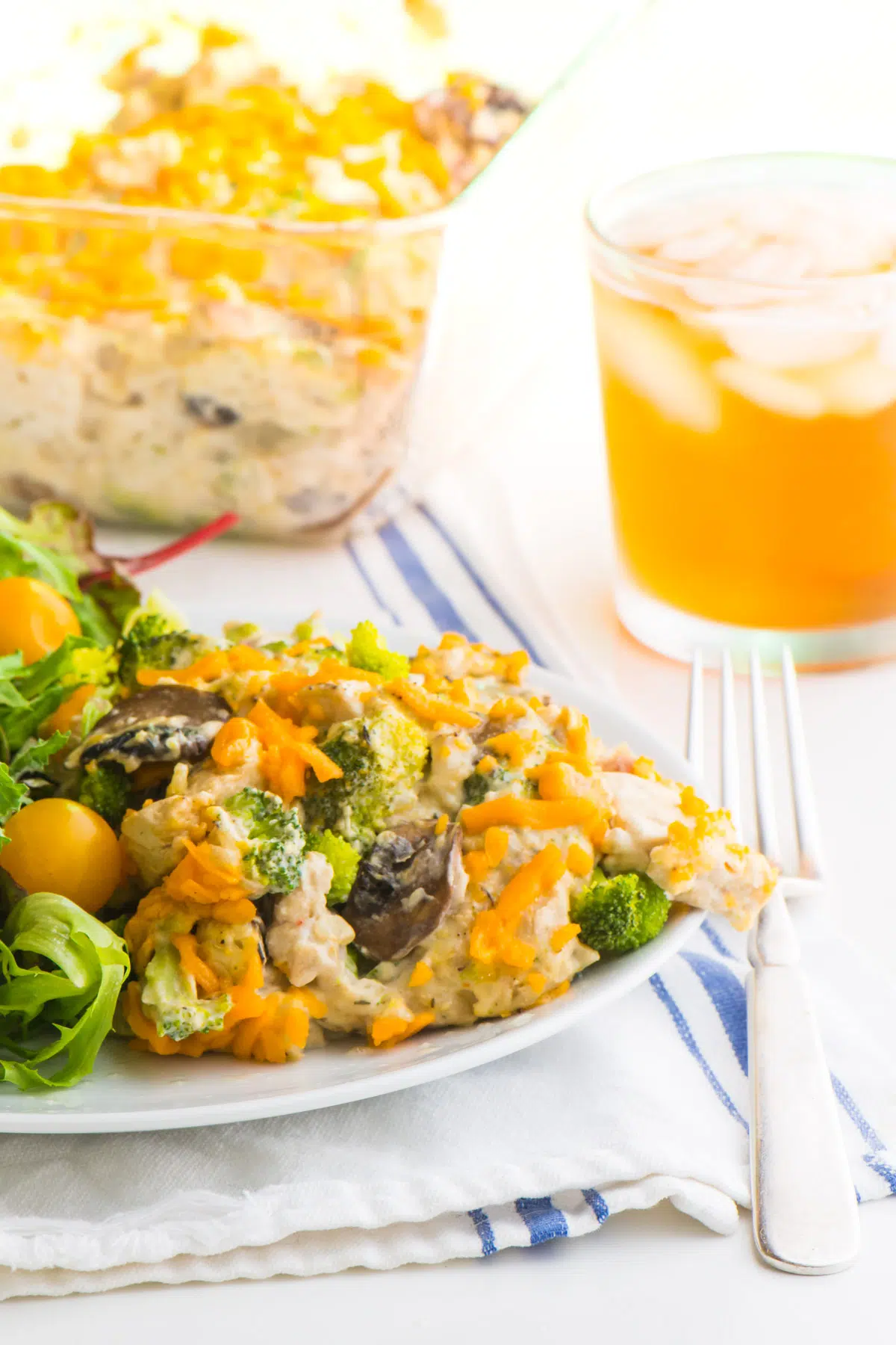A serving of vegan chicken and rice casserole on a plate with a green salad. A fork is sitting next to it, with a glass of iced tea and the rest of the casserole behind it.