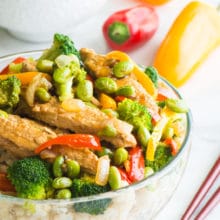 A vegan teriyaki chicken and edamame rice bowl sits next to chopsticks with a bowl of rice behind it and mini bell peppers.