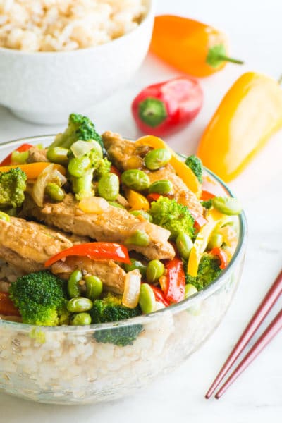 A vegan teriyaki chicken and edamame rice bowl sits next to chopsticks with a bowl of rice behind it and mini bell peppers.