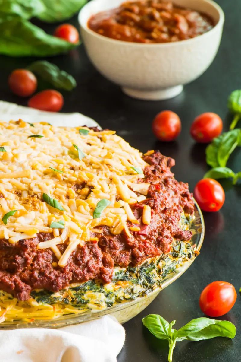 A pie plate full of vegan spaghetti pie with melted vegan cheese on top sits on a dark countertop. There are fresh cherry tomatoes sitting around the the dish, with fresh basil and a bowl full of pasta sauce in the background.