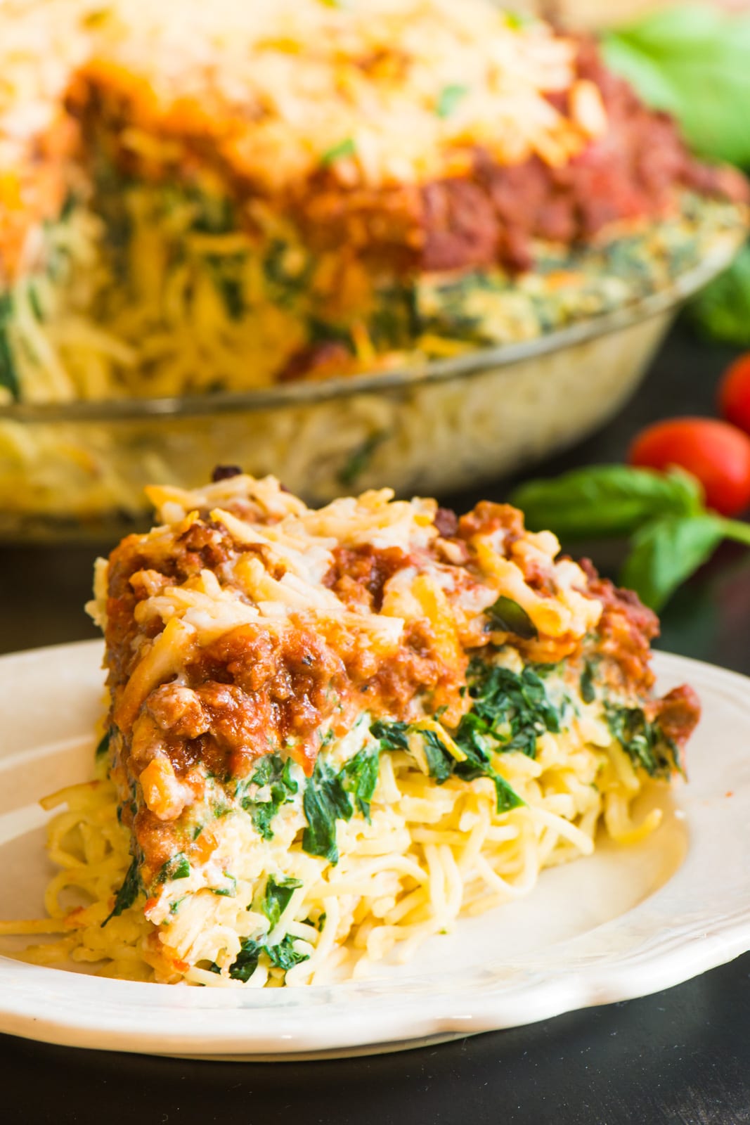 A slice of vegan spaghetti pie sits on a plate. You can see layers of spaghetti, spinach, red sauce, and melted cheese on the top. Behind it is the entire dish with a slice removed. There's fresh basil and tomatoes beside it.