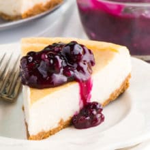 A slice of vegan New York style cheesecake sits on a plate with blueberry sauce over the top.