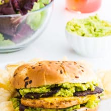 A Vegan Chicken Sandwich with Avocados sits on a plate surrounded by potato chips with a bowl of guacamole and bottle in the back