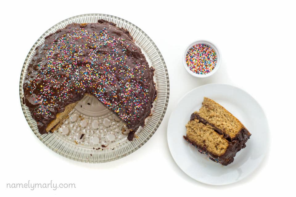 Looking down on a slice of vegan vanilla wacky cake on a plate beside the whole cake with a few slices cut out. A bowl of sprinkles sits beside the plate.