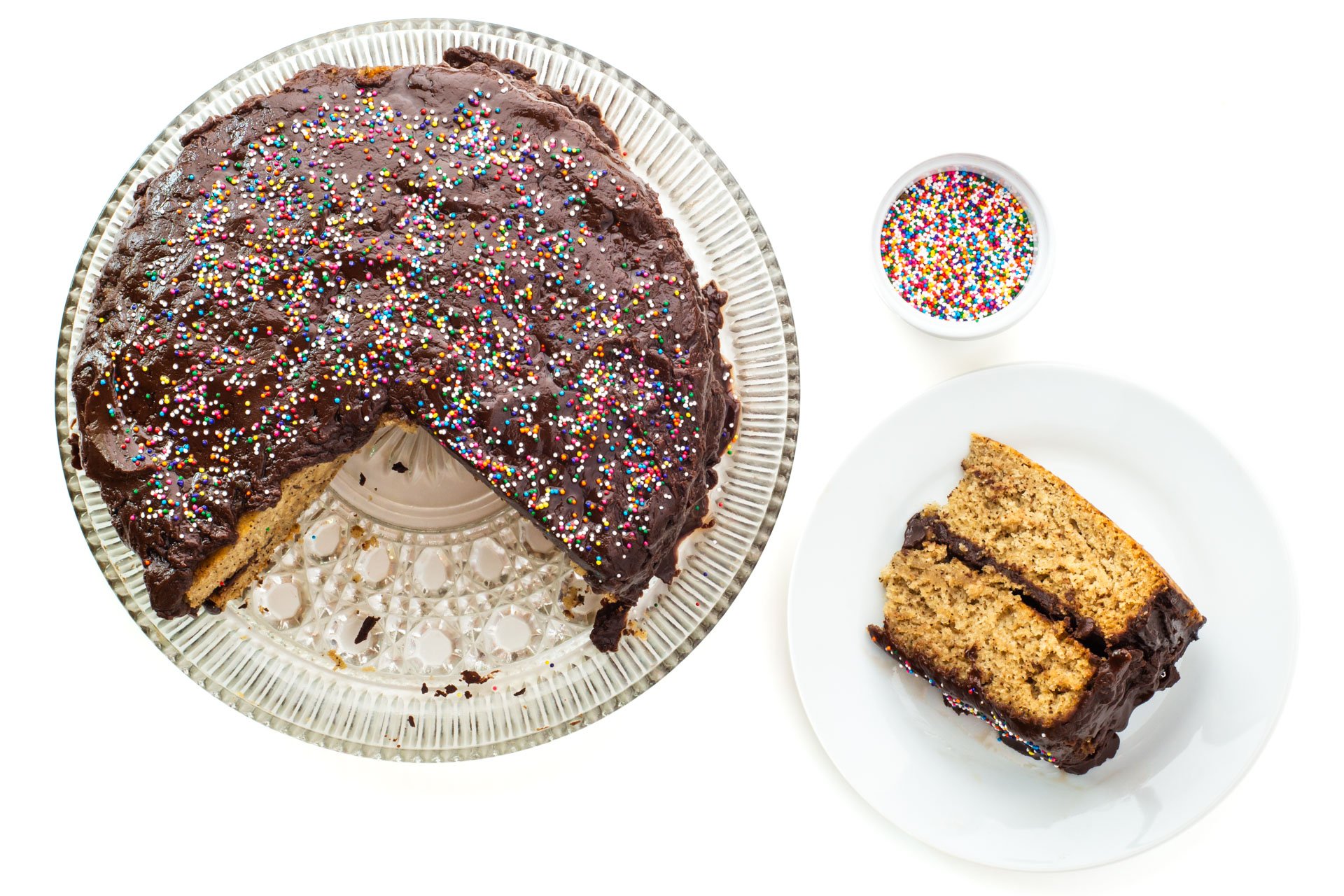 Looking down on a slice of vegan vanilla wacky cake on a plate beside the whole cake with a few slices cut out. A bowl of sprinkles sits beside the plate.