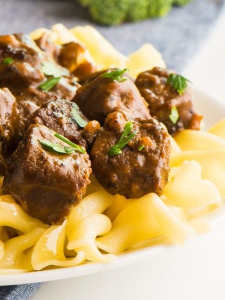 A white plate topped with vegan beef stroganoff shows noodles and parsley sprinkled over the vegan beef pieces. There is steamed broccoli in the background.
