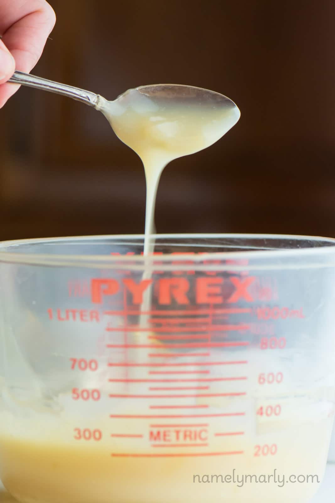 A spoon drizzles a creamy milk mixture over a pyrex measuring cup.