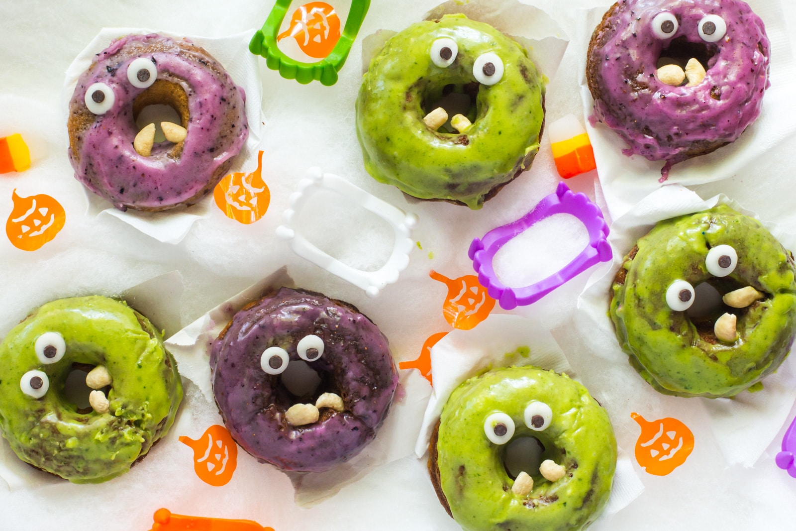Several vegan halloween monster donuts with plastic vampire teeth and orange pumpkins and candy corn between them.
