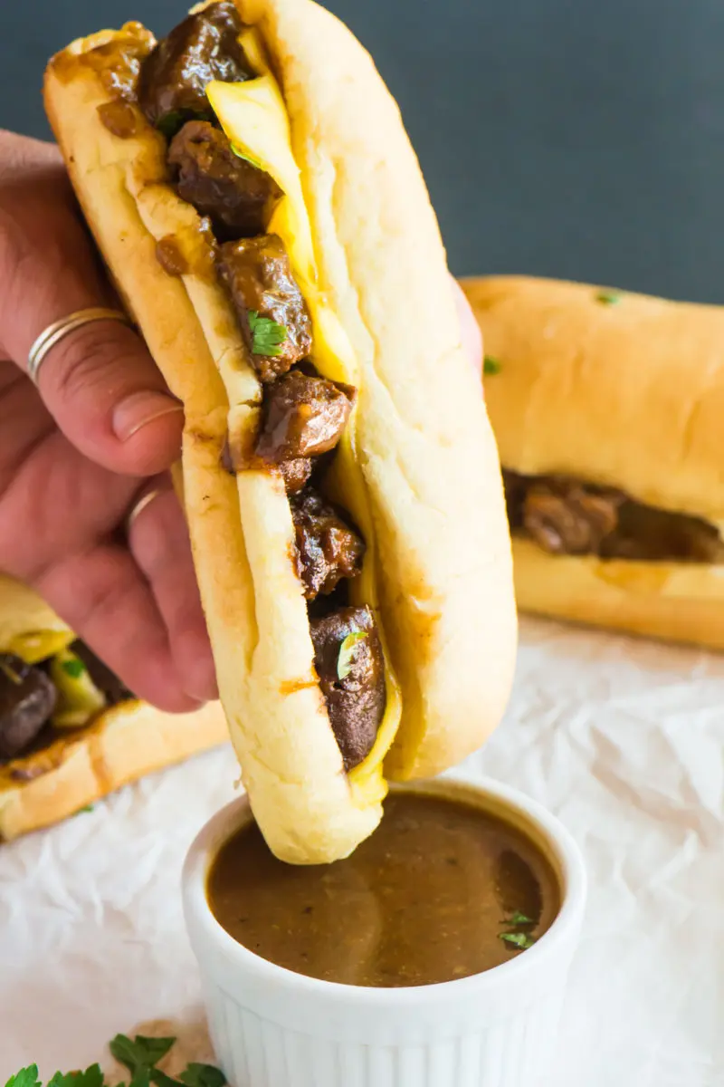 A hand is dipiping a Vegan French Dip Sandwich into a small bowl of sauce.