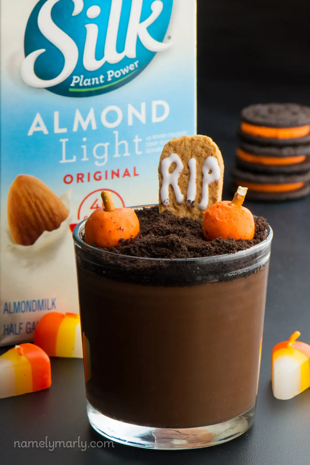 A vegan Halloween pudding cup has candy pumpkins and a graham cracker that reads, "RIP" and sits in front of orange filled Oreos and a carton of plant-based milk used to make it.