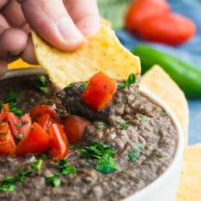 A hand holds a tortilla chip with black bean dip on it over the rest of the dip in a bowl.