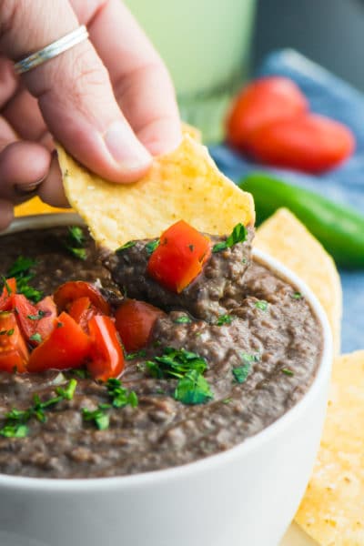 A hand holds a tortilla chip with black bean dip on it over the rest of the dip in a bowl.