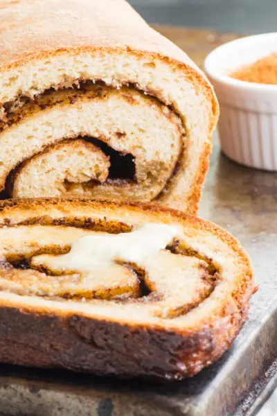 A slice of vegan cinnamon bread with melted vegan butter sits next to the rest of the loaf.