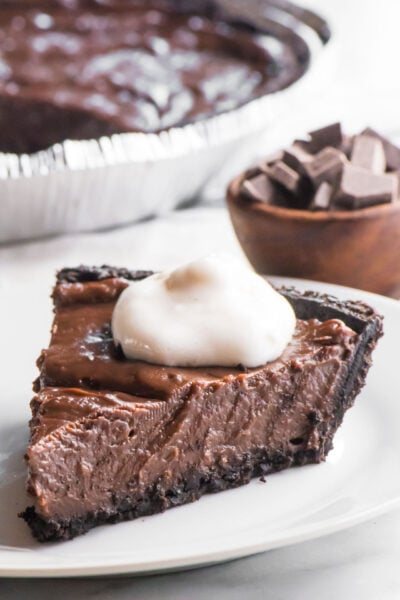A slice of vegan peanut butter pie sits on a plate. There's a bowl of chocolate sauce behind it along with the rest of the pie.