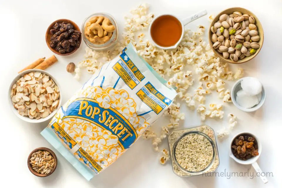 Several ingredients are on a white counter, including a bag of popped microwave-popcorn, and bowls of nuts, raisins, syrup, coconut oil, and more.