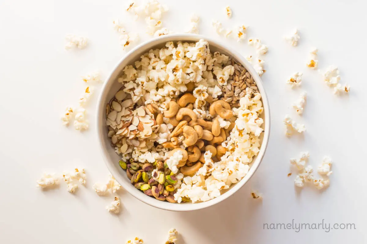 Popcorn is in a large bowl with nuts and seeds. There re popped popcorn kernels around the bowl.