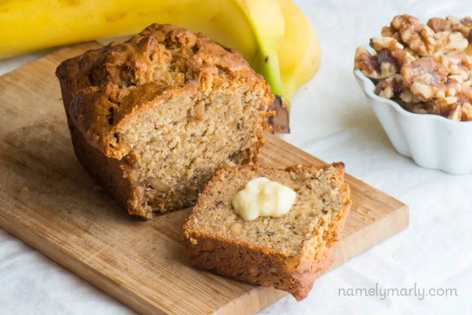 A slice of quick bread with melted butter sits next to the rest of the loaf with a bowl of walnuts beside it and a couple of bananas.