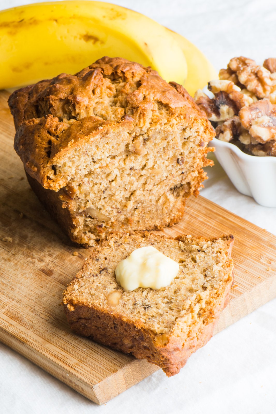 A slice of vegan banana nut bread is topped with melted vegan butter with the rest of the loaf behind it.