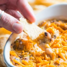 A hand holds a tortilla chip full of some vegan black-eyed pea dip in front of the rest of the dip.