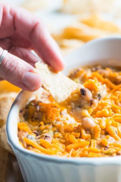 A hand holds a tortilla chip full of some vegan black-eyed pea dip in front of the rest of the dip.