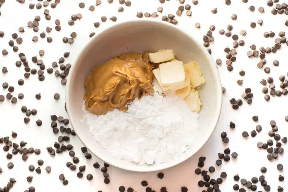 A bowl has powdered sugar, vegan butter, and peanut butter inside it. It is surrounded by scattered chocolate chips.
