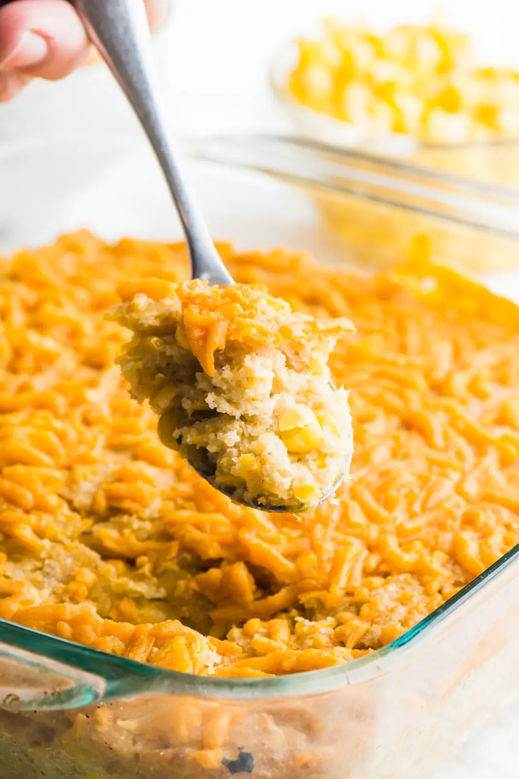 A spoonful of vegan corn casserole is hovering over the entire dish, with a bowl of corn behind it.
