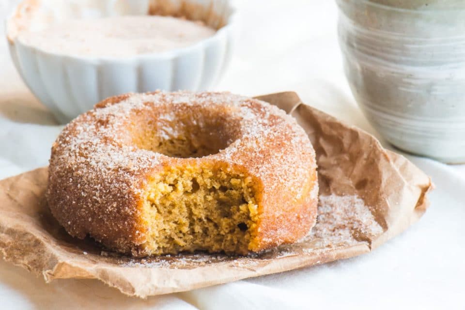 A pumpkin donut with a bite taken out sits on brown paper. A bowl of cinnamon sugar sits behind it.