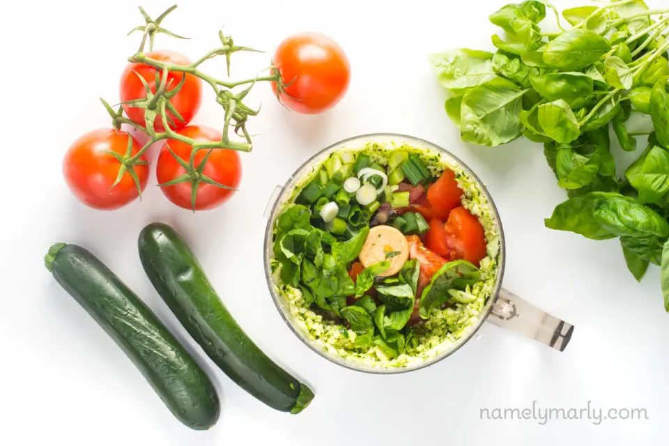 Looking down on ingredients in a food processor next to spinach, zucchini, and tomatoes on the vine.