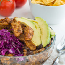 A vegan enchilada bowl, topped with avocados, red cabbage and served with a bowl of tortilla chips and fresh tomatoes in the background.