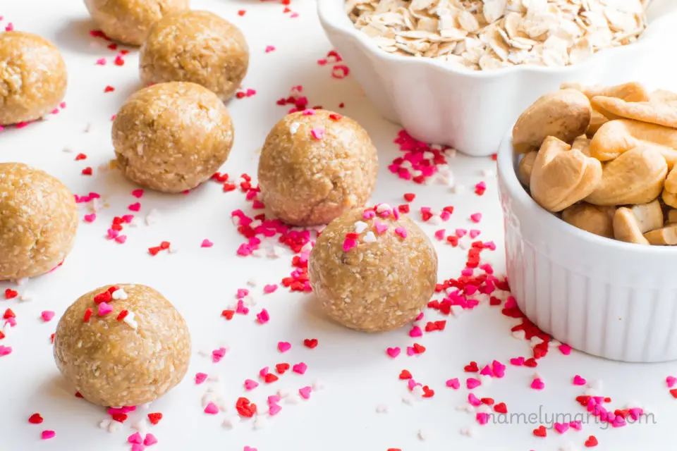 Several no-bake sugar cookie energy bites sit on a counter top with a bunch of multi-colored heart-shaped sprinkles around them. There are two white bowls holding ingredients, including whole cashews and rolled oatmeal.