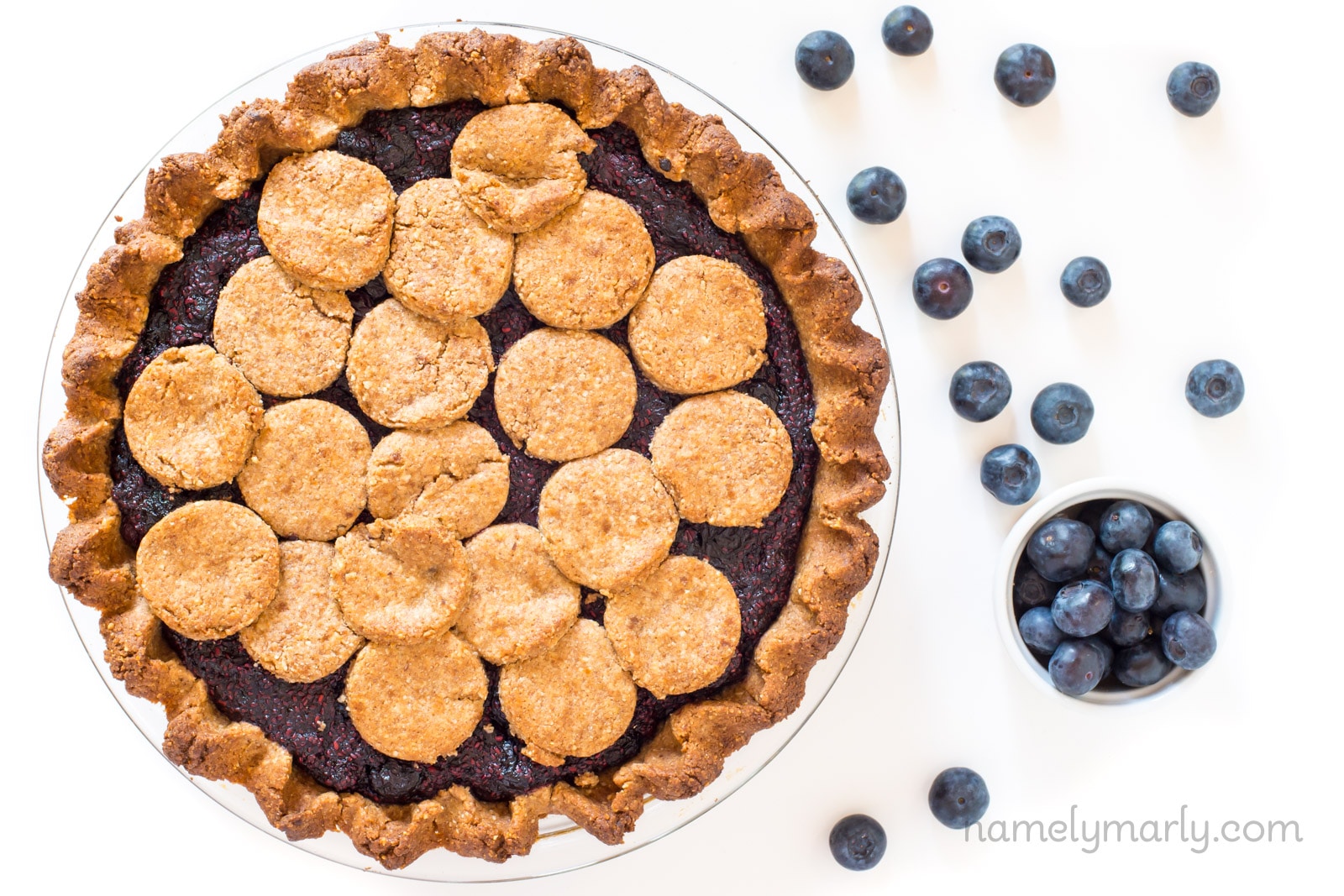 An overhead view of a berry pie with blueberries scattered around on a marble surface