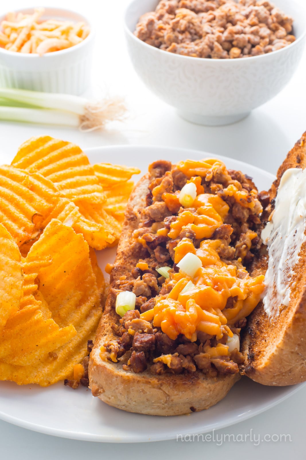 A vegan chopped cheese sandwich sits on a plate next to potato chips with ingredients in the background to make more.