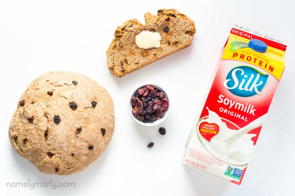 A round loaf of vegan Irish soda bread sits next to a bowl of dried cranberries, a slice of the bread, and a carton of soy milk.