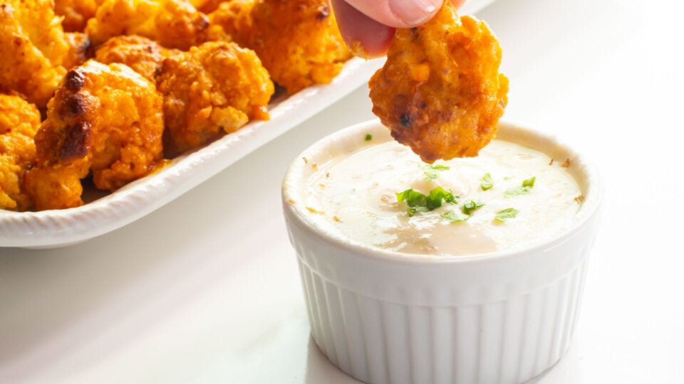 A hand dips Vegan Baked Cauliflower Wings into a bowl of ranch dressing.