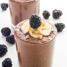 A serving glass holds vegan chocolate chia pudding topped with sliced bananas and fresh blackberries. Another glass and more blackberries is behind it.
