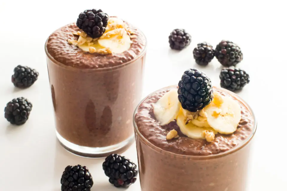A closeup of two serving glasses full of chocolate chia pudding, topped with sliced bananas and fresh blackberries.