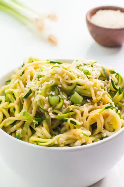 A bowl of spicy sesame zoodles with sesame seeds sits in front of green onions and a bowl of more sesame seeds.