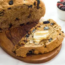 A loaf with raisins has a slice cut out with melted vegan butter and a bowl of raisins beside it.