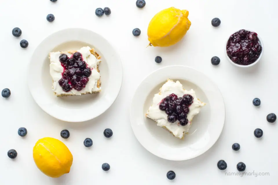 Looking down on two slices of cake surrounded by lemons, blueberries, and blueberry sauce in a bowl.