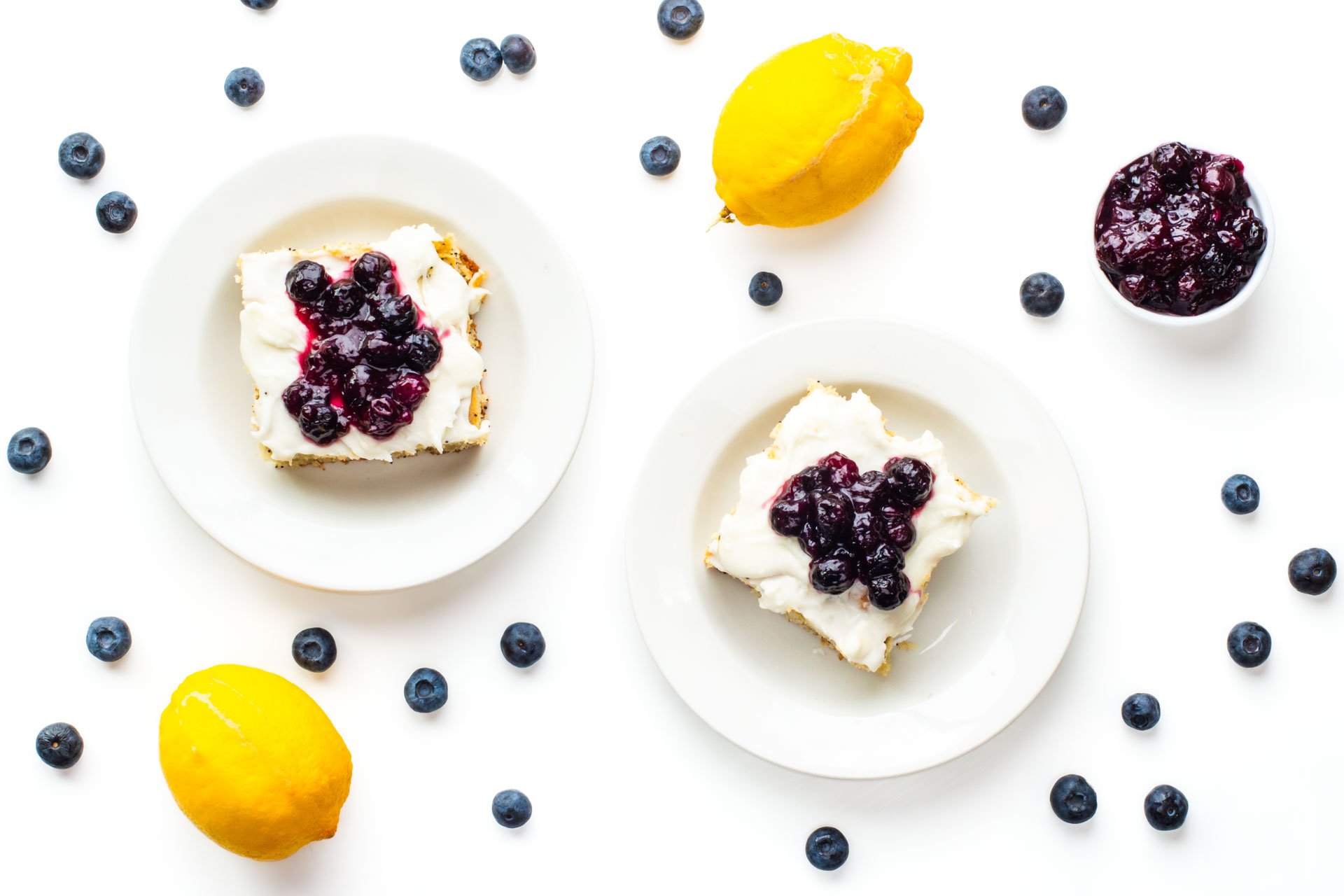 Looking down on two slices of cake surrounded by lemons, blueberries, and blueberry sauce in a bowl.