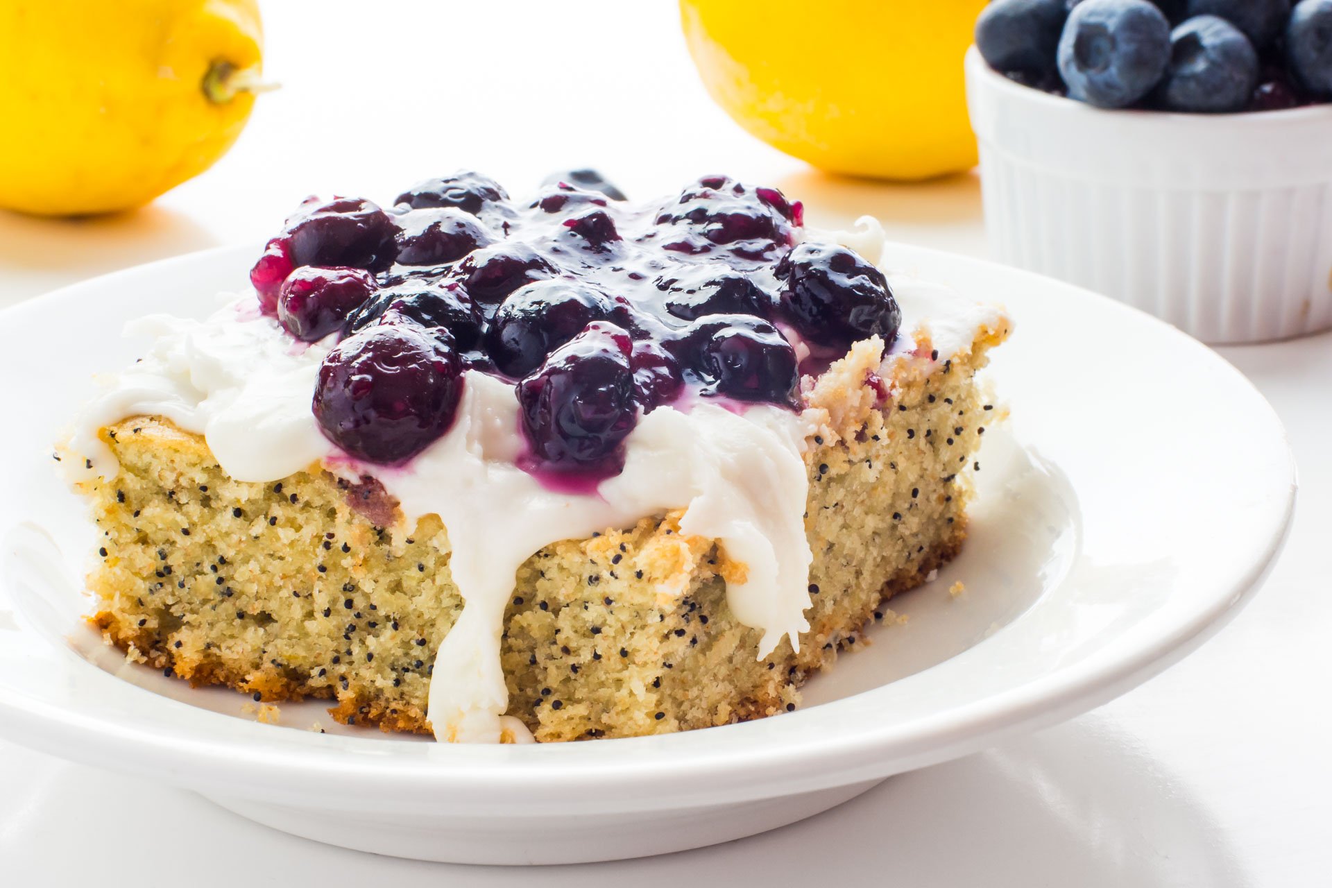 A slice of Lemon Avocado Cake with Blueberry Topping sits in front of lemons and fresh blueberries.