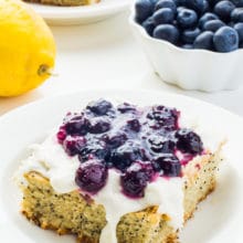 A slice of lemon avocado cake with blueberry sauce on top sits on a plate near lemons, a bowl of blueberries and another slice of cake.