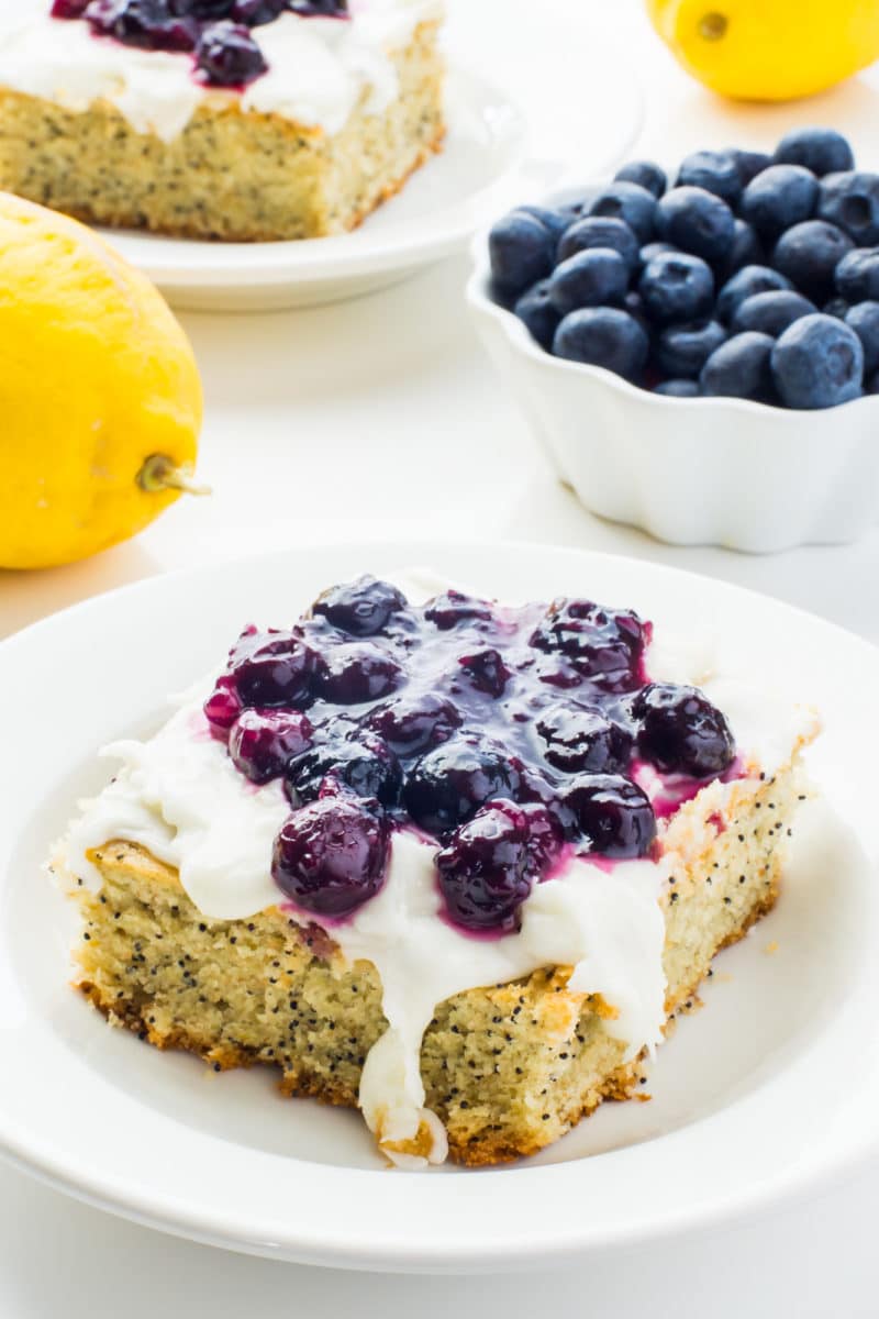 A slice of lemon avocado cake with blueberry sauce on top sits on a plate near lemons, a bowl of blueberries and another slice of cake.