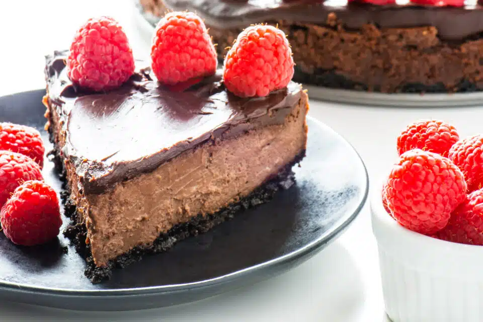 A slice of vegan chocolate cheesecake sits on a black plate with fresh raspberries on top and in a bowl beside it.