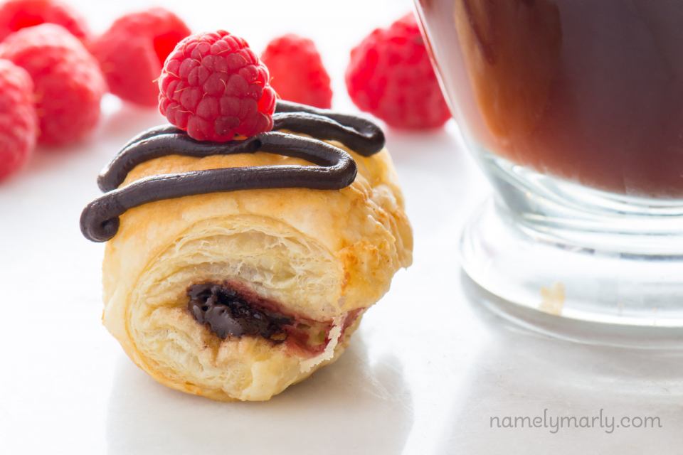 A Vegan Chocolate Croissants with chocolate drizzle and a red raspberry on top sits beside a glass mug of coffee.