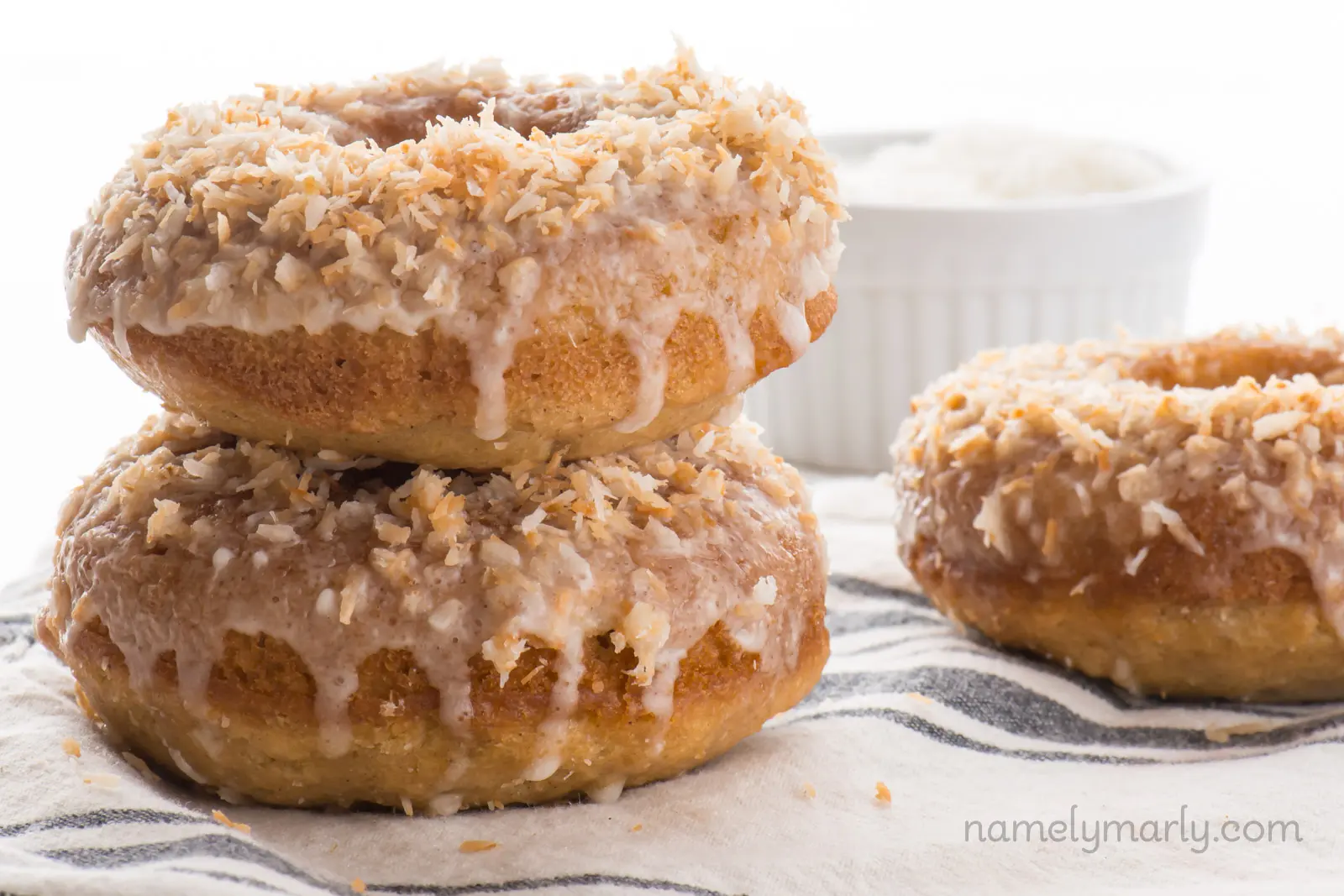 A stack of two Vegan Baked Toasted Coconut Donuts is sitting beside another donut, with a bowl of coconut flakes behind it.
