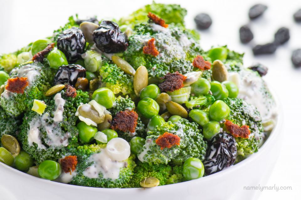 A close-up shot of Vegan Broccoli Salad with peas, pumpkin seeds, and veggie bacon pieces featured. There are additional raisins in the background.