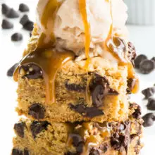 A stack of vegan caramel chocolate chip cookie bars with vanilla ice cream on top and melted caramel drizzling over the sides.