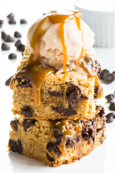 A stack of vegan caramel chocolate chip cookie bars with vanilla ice cream on top and melted caramel drizzling over the sides.